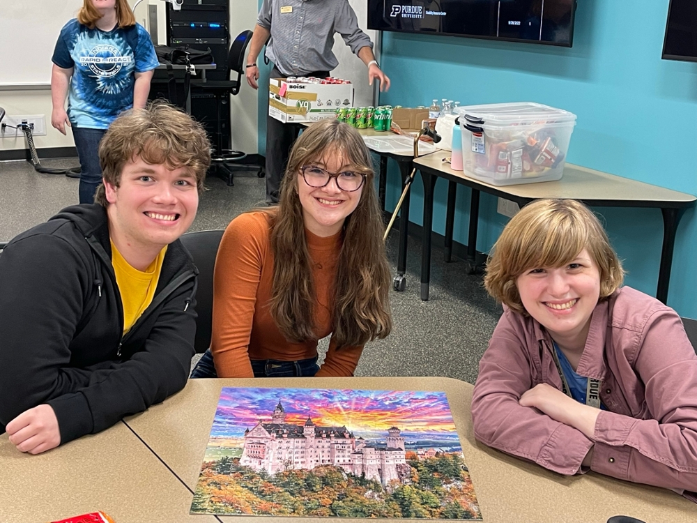 Pictured: three students sit around a puzzle during a Peer Mentor Program game night, smiling at the camera