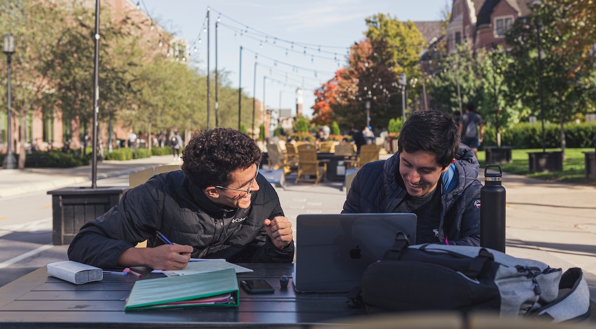 Pictured: two students sit outside of krach leadership center talking and working on laptops