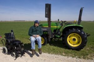 Farmer sitting on lift to raise him into tractor seat while wheelchair sits on left of photo