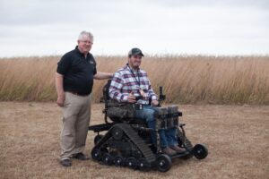 Man sits in heavy-duty tracked wheelchair while other man stands next to him with tall grain in field behind them.