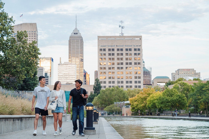 Picture of three college-aged students walking next to a canal with downtown Indianapolis buildings in the background.