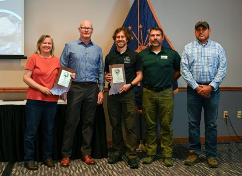 Five individuals stand indoors, three holding certificates, with an Indiana state flag in the background during an award ceremony.