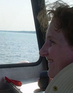 A person behind the wheel of a boat. They are smiling and there is blue water on the horizon.
