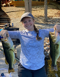 A person in a blue shirt wearing a baseball hat, holding up a fish in each hand. There are trees and a pathway behind them.