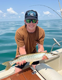 A person in a brown shirt on the side of a boat. They are holding a fish in both hands. There is a fishing rod to the side, and blue water and sky behind them.