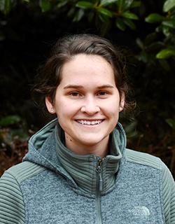 A person in a green coat. They are smiling. There are dark green leaves in the background.