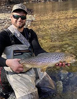 A person in a baseball hat and chest waders holds a fish horizontally in both hands. They are wearing sunglasses and smiling. There is water behind them.