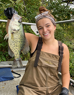 A person wearing chest waders holds up a fish. They are on a boat and they are smiling. There are green trees in the background.