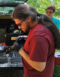 A person in a dark red shirt holding tools. There is a pickup truck behind them. A hand is holding something up to the left, and a person in a bright blue shirt is behind them.