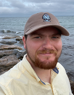 A person wearing a baseball hat and a yellow shirt. They have a beard and mustache. There are rocks and water in the background.