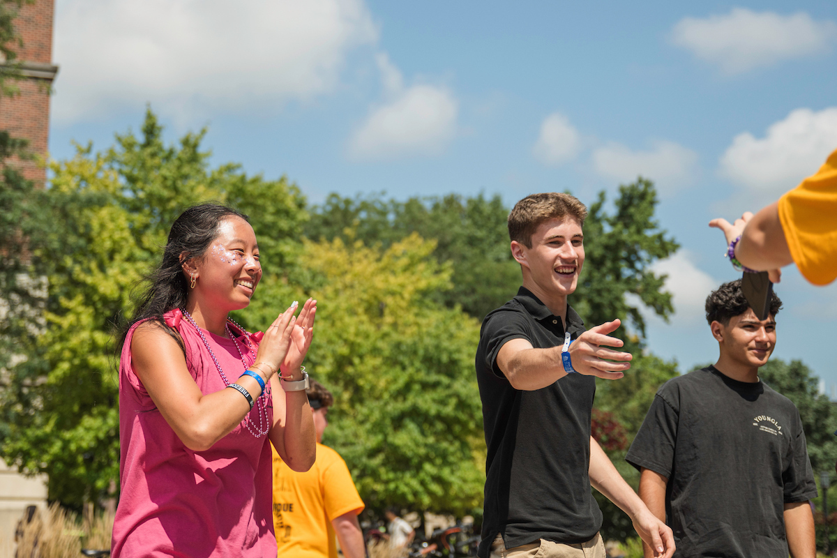 A group of young people outdoors, smiling and clapping under a sunny sky with trees and a brick building in the background.