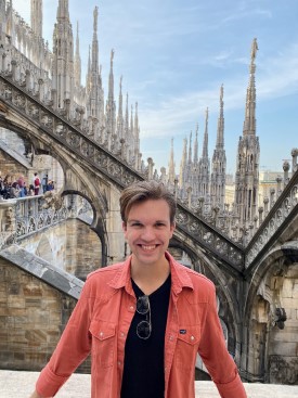 Andrew Arndt wears a red shirt and stands on the roof of the Milan Duomo with spires and ornate stonework in the background