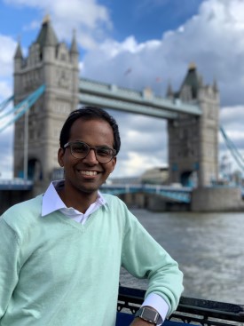 Arvindakshan Rajesh, wearing a collared shirt and glasses, smiles in front of London's Tower Bridge