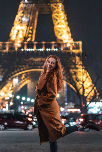 Carmen Michlovich poses and pops her foot up in front of the Eiffel Tower light up in sparkling lights at night