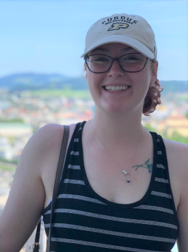 Joanna Bastian wearing a white Purdue baseball cap and striped tank top smiles in front of a city scence