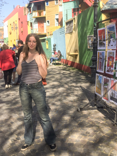 Victoria Malone stands in front of bright and colorful houses in Buenos Aires, with a vendor selling art in the background