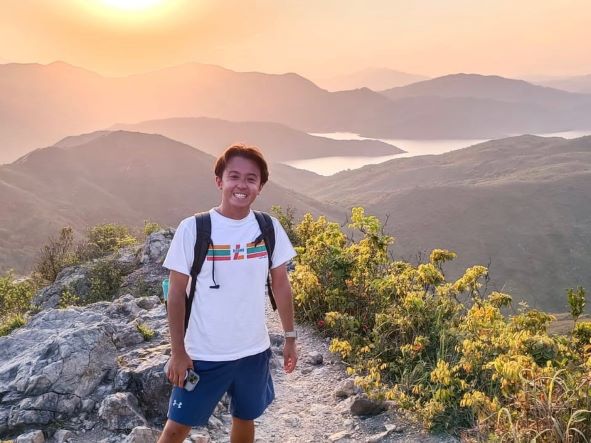 Nick Mori stands on a rocky landscape with mountains and a hazy sunset in the distance
