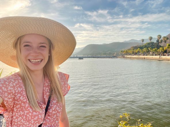 Rachael Snow smiles wearing a big straw hat with a sunny scene of a lake in the background