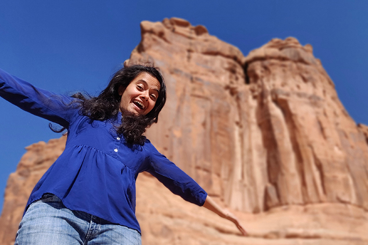 Deepti Tagare smiling with her arms open, outside, in front of a stone feature