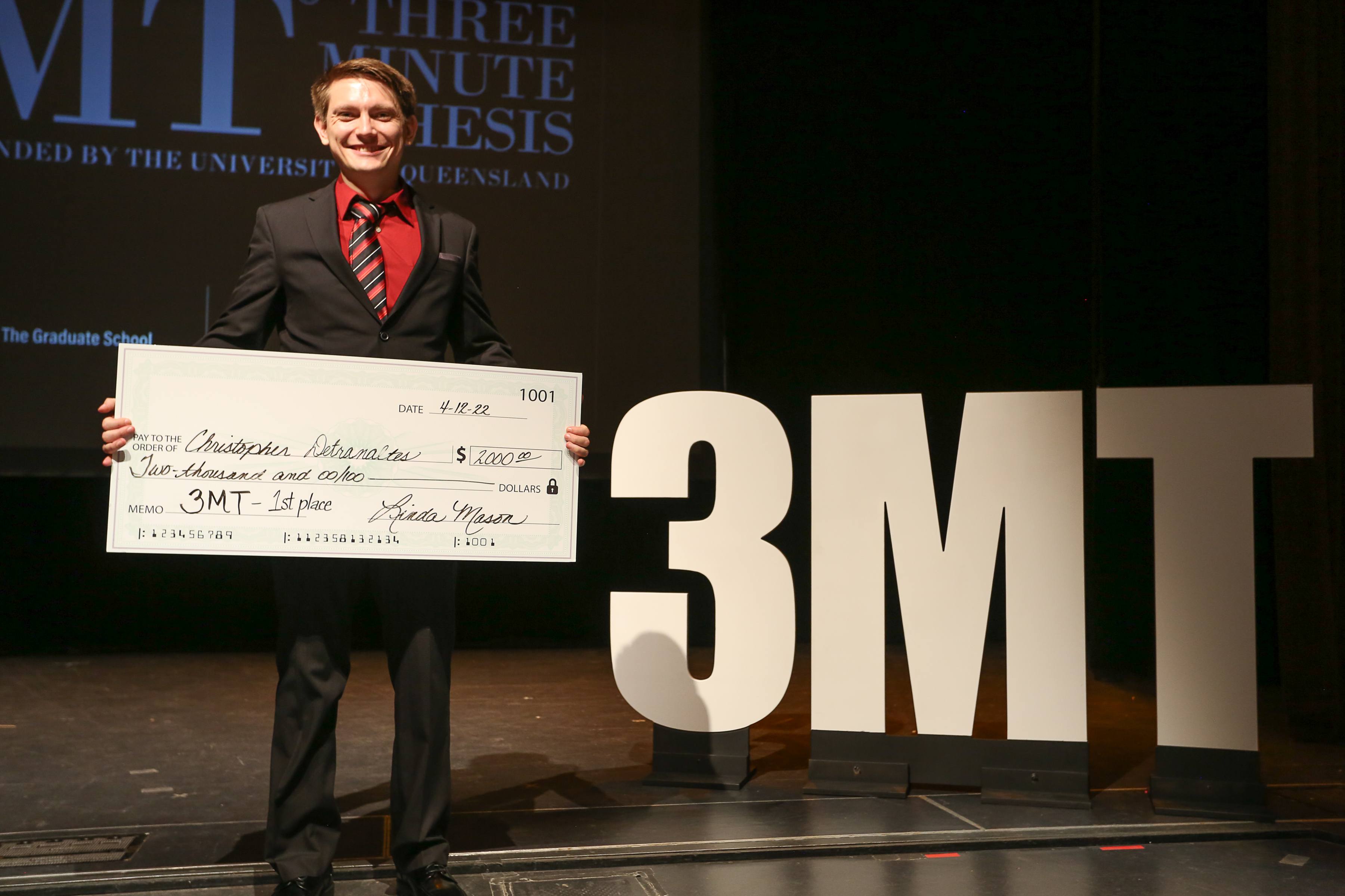Student posing on stage by the 3MT logo with a first prize check