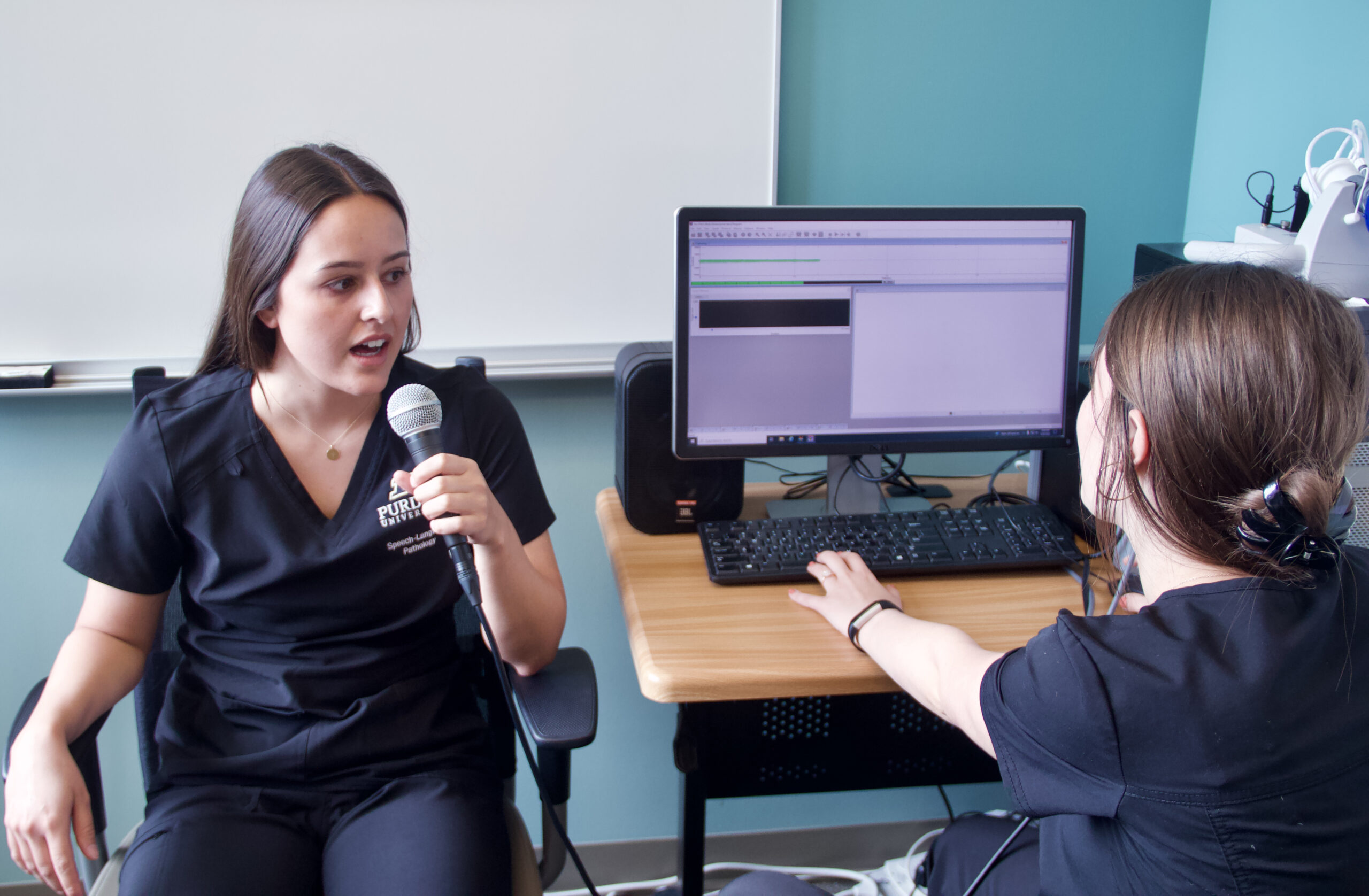 Savannah Limcaco performs a vocal exercise into a microphone connected to a computer while Mikahla Combs measures the output.