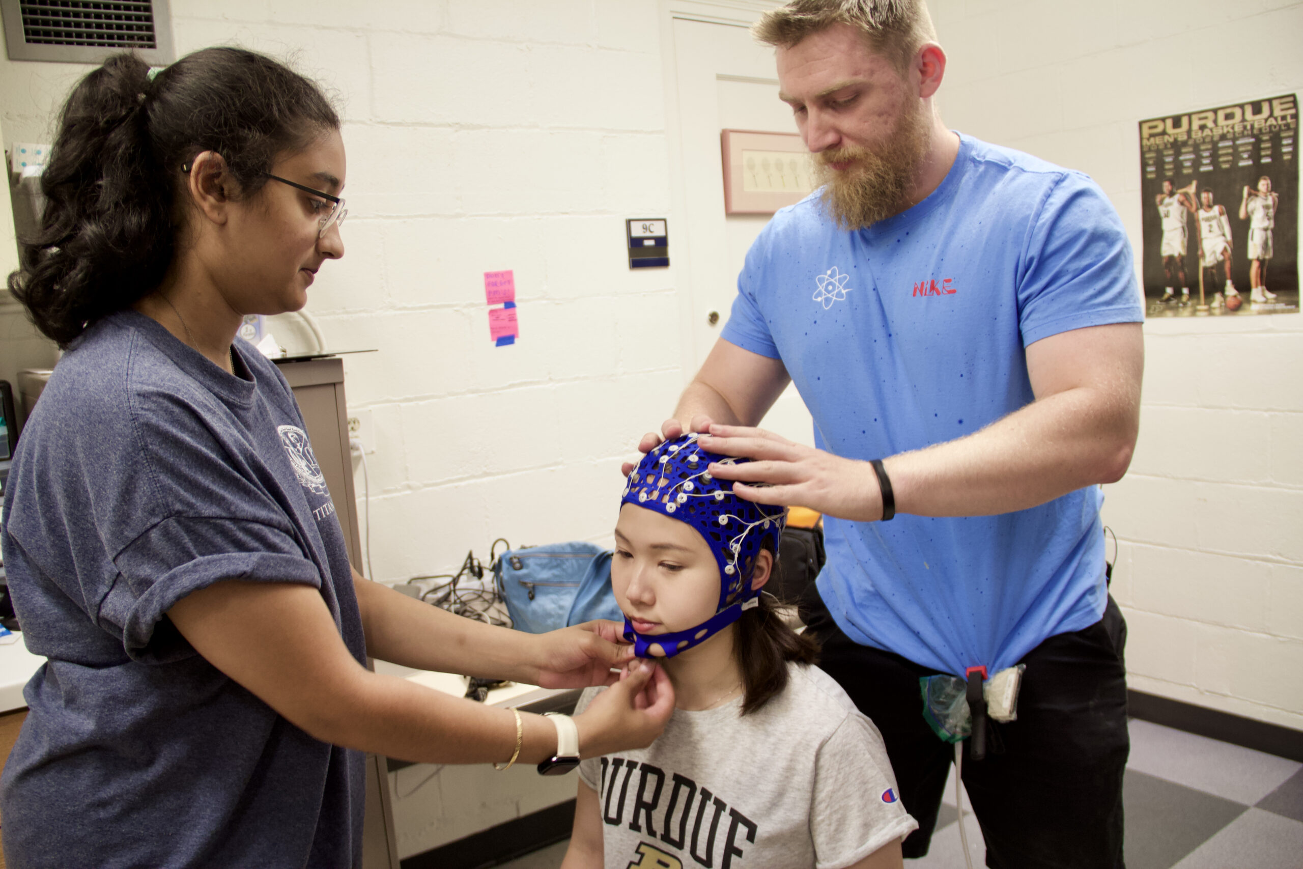 Rida Khatri and Nick Baumgartner stand to fit an EEG cap to Amber Yu, who is seated.