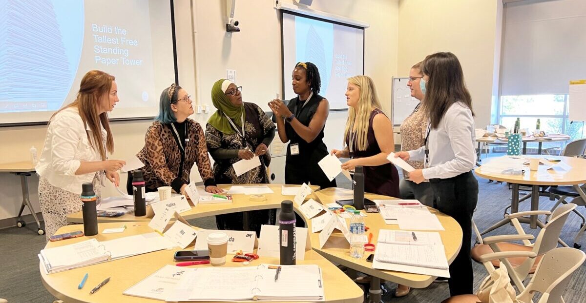 Seven women gather around a table working on a project creating paper towers.