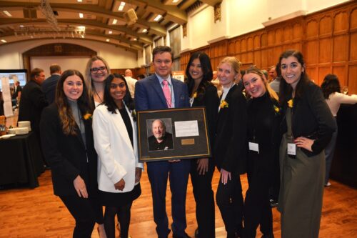 Seven women and one man pose with a plaque in a networking reception.