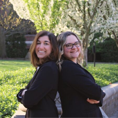 Libby Loprete and Madi Lindsey stand back-to-back in business attire in a grassy area