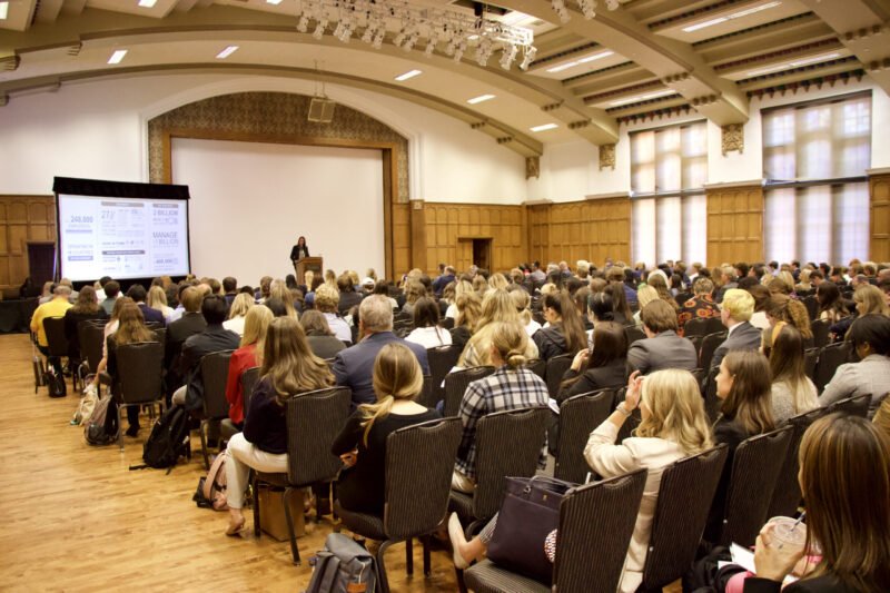 People sit in rows in a full room listening to a speaker at a podium.