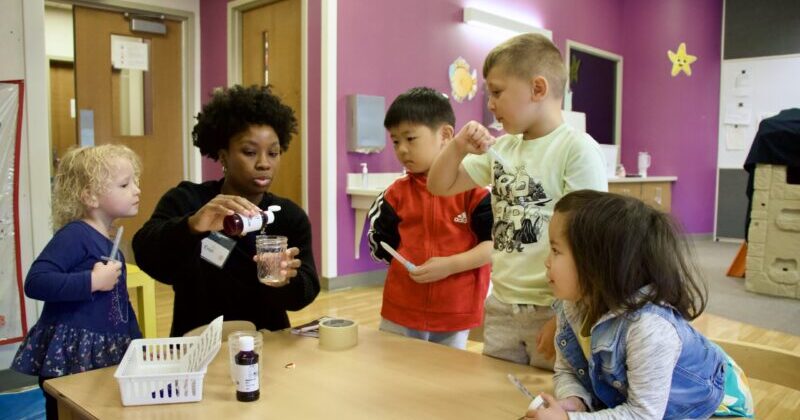 A woman sits at a table with four children working on a craft