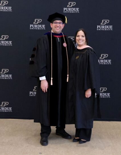 A man and a woman stand in graduation gowns, smiling.