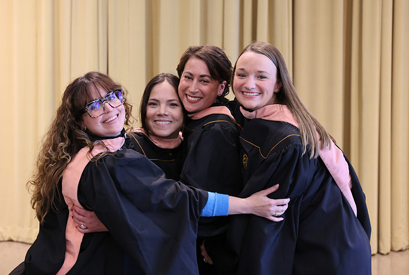 Four women in graduation gowns hug and smile for a photo.