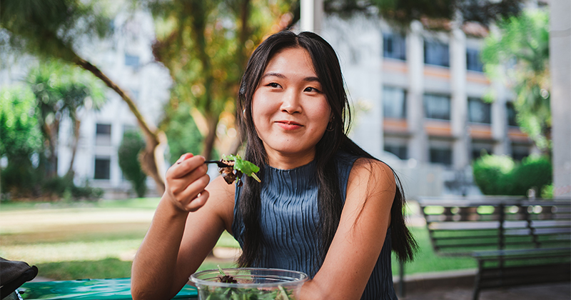 A girl sits at a table and eats a salad, smiling