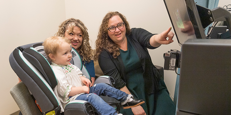 A woman stands next to a mom and child and points to a monitor attached to the wall.