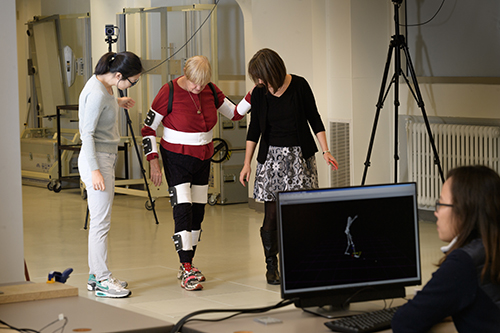 An older woman walks with monitors on her body to track her movement.