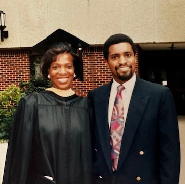 Two smiling people pose together, one in a graduation gown.