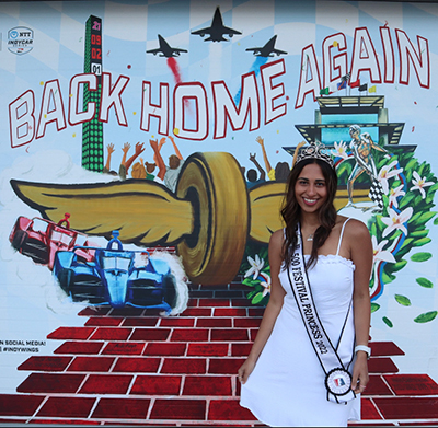 Emily Deldar poses in a crown and dress in front of a sign that reads "Back Home Again" with Indy 500 cars on it.
