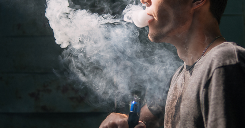 A younger man's profile is seen holding a vaping device with a cloud of smoke in front of him.