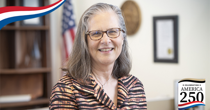 Shelley MacDermid Wadsworth stands in her office. An America250 graphic appears in the bottom righthand corner.
