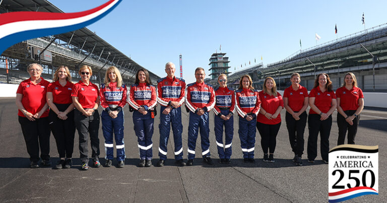 A group of medical team members dressed in red and black outfits line up along the Indianapolis Motor Speedway track for a group picture. An America250 graphic appears in the bottom right corner.