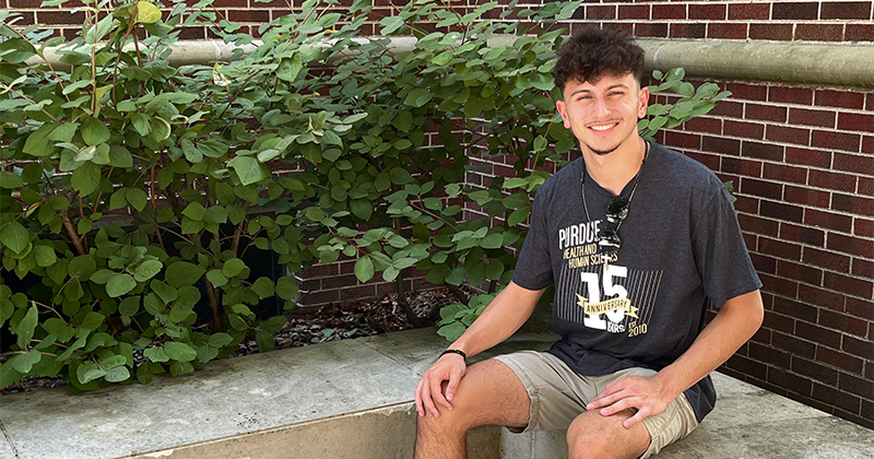 Preston Serratos sits in front of a brick building with greenery, smiling