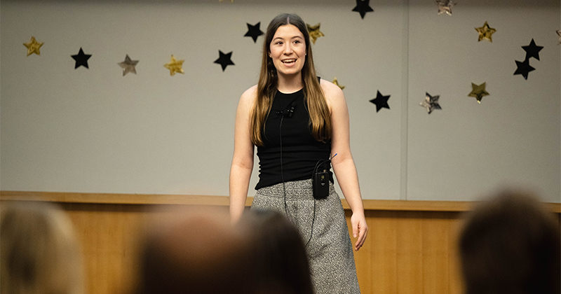 A young woman stands in front of a crowd with a microphone, speaking.