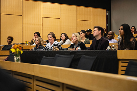People sit in a lecture hall, smiling and listening to a speaker.