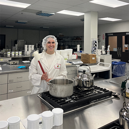 A woman stands in a white coat and hair net by a large pot, holding a spatula.