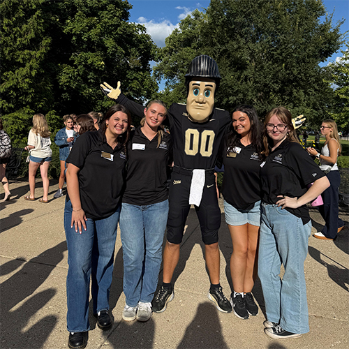 A group of four girls in matching black polos pose for a photo with Purdue Pete.