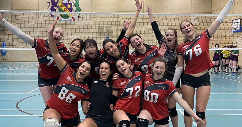 A group of girls in volleyball uniforms stand in front of a volleyball net, cheering.