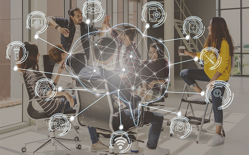 A group of working professionals sit together at a conference table, talking. A graphic of a brain with tech symbols sits on top of the image, slightly transparent.