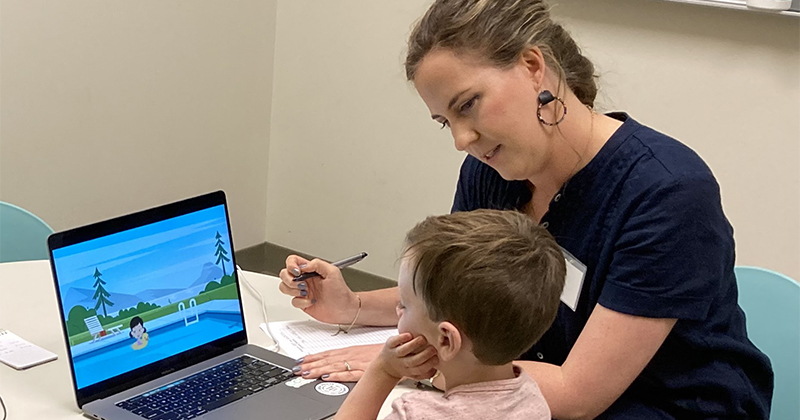 A child looks at a picture on a laptop screen with a woman taking notes on a piece of paper, observing him.