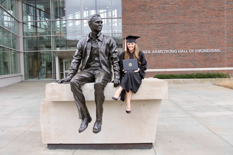 Taylor sits next to the Neil Armstrong sculpture outside wearing her graduation cap and gown.
